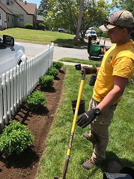A man in a yellow shirt is standing in front of a white picket fence holding a shovel.