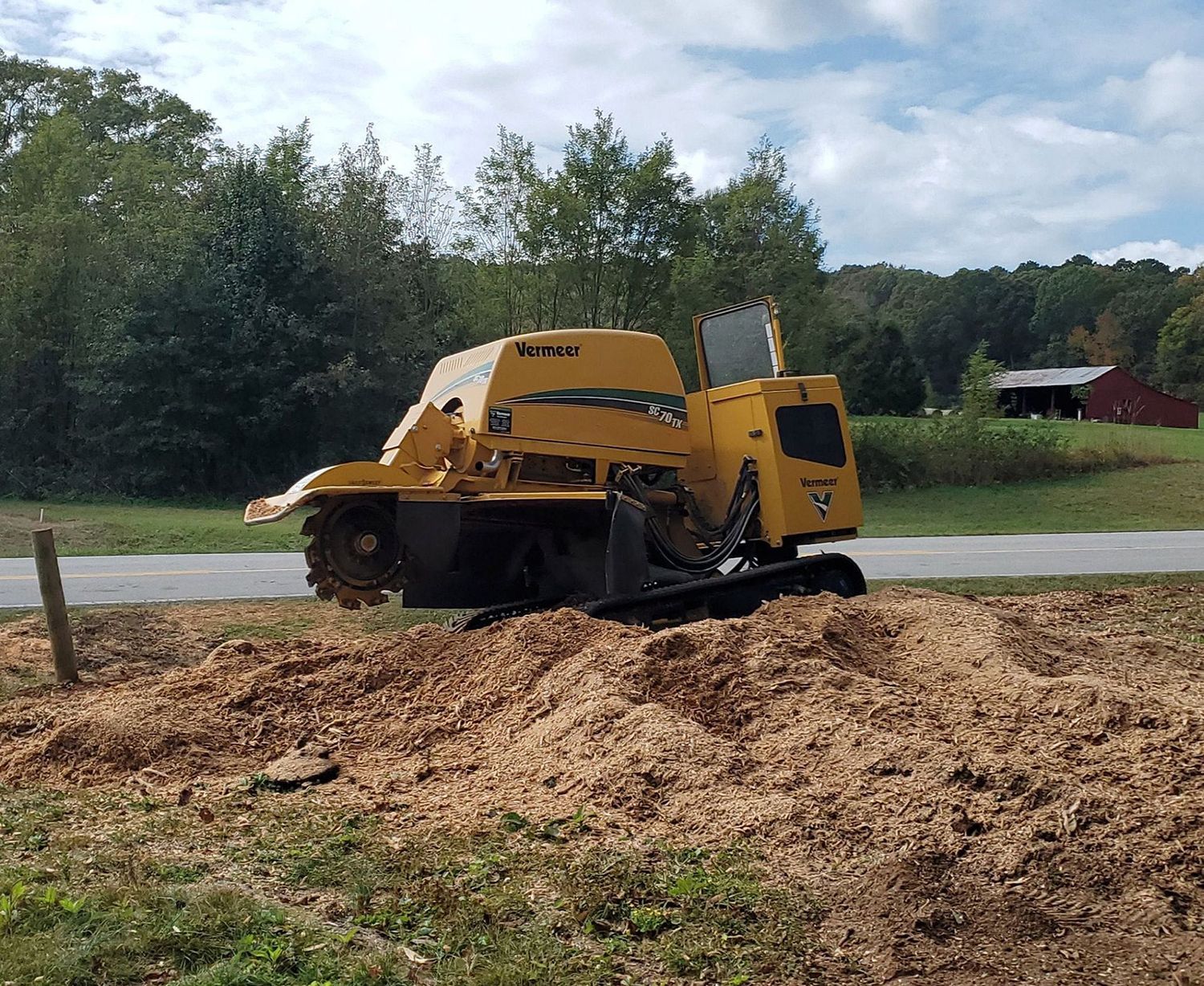 A yellow industrial wood chipper sits on a large pile of wood chips in a field under a partly cloudy blue sky.