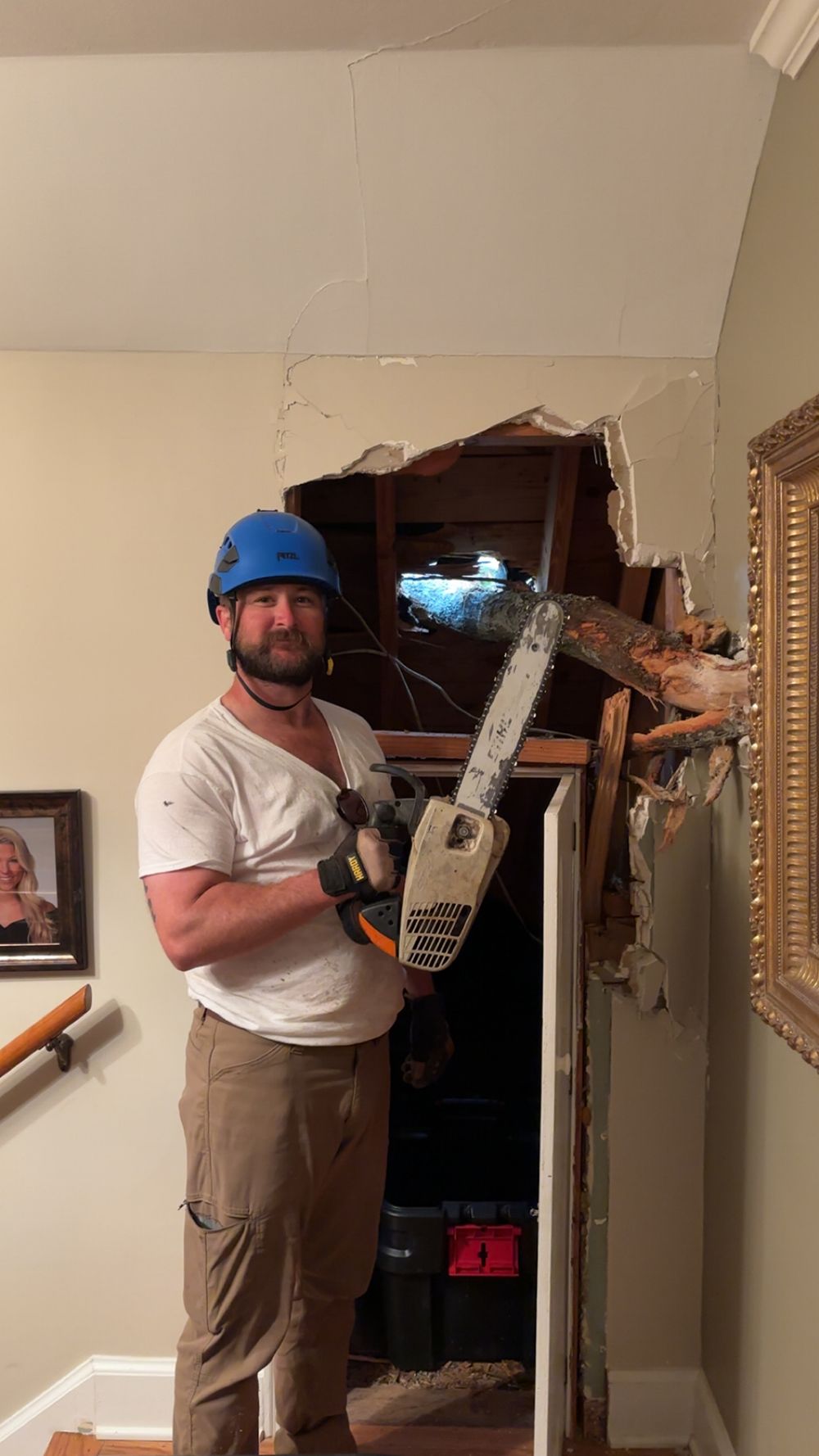 A person in a blue hard hat holding a chainsaw stands next to a hole they have cut in an interior wall.