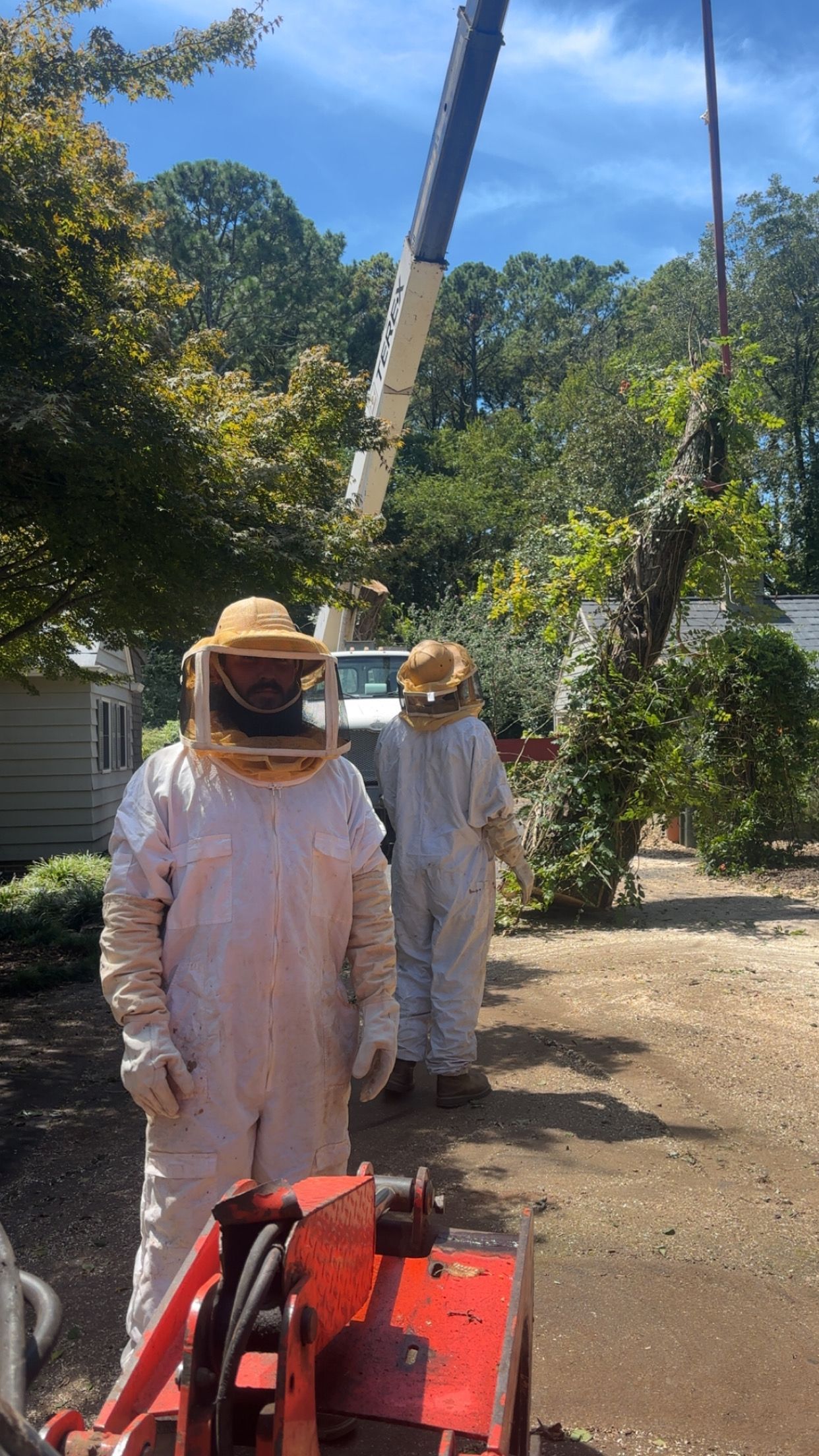 Two people wearing white protective beekeeping suits and veils stand outdoors near a crane and a tree.