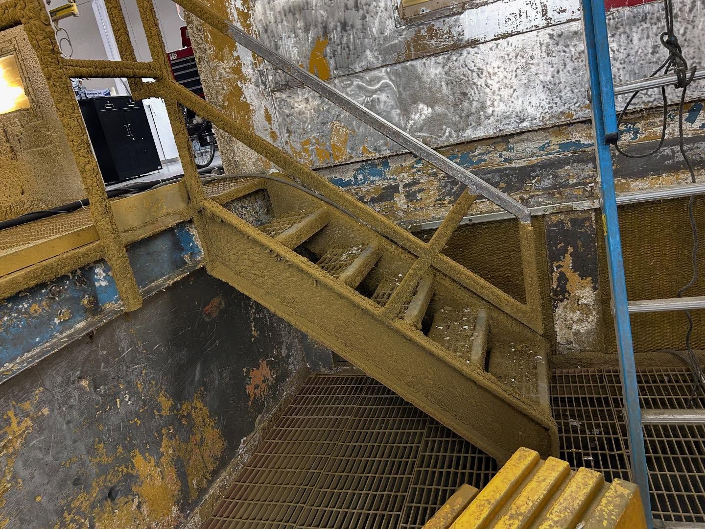 Wooden staircase covered in yellow paint, leading to an upper level. Grated metal floor.