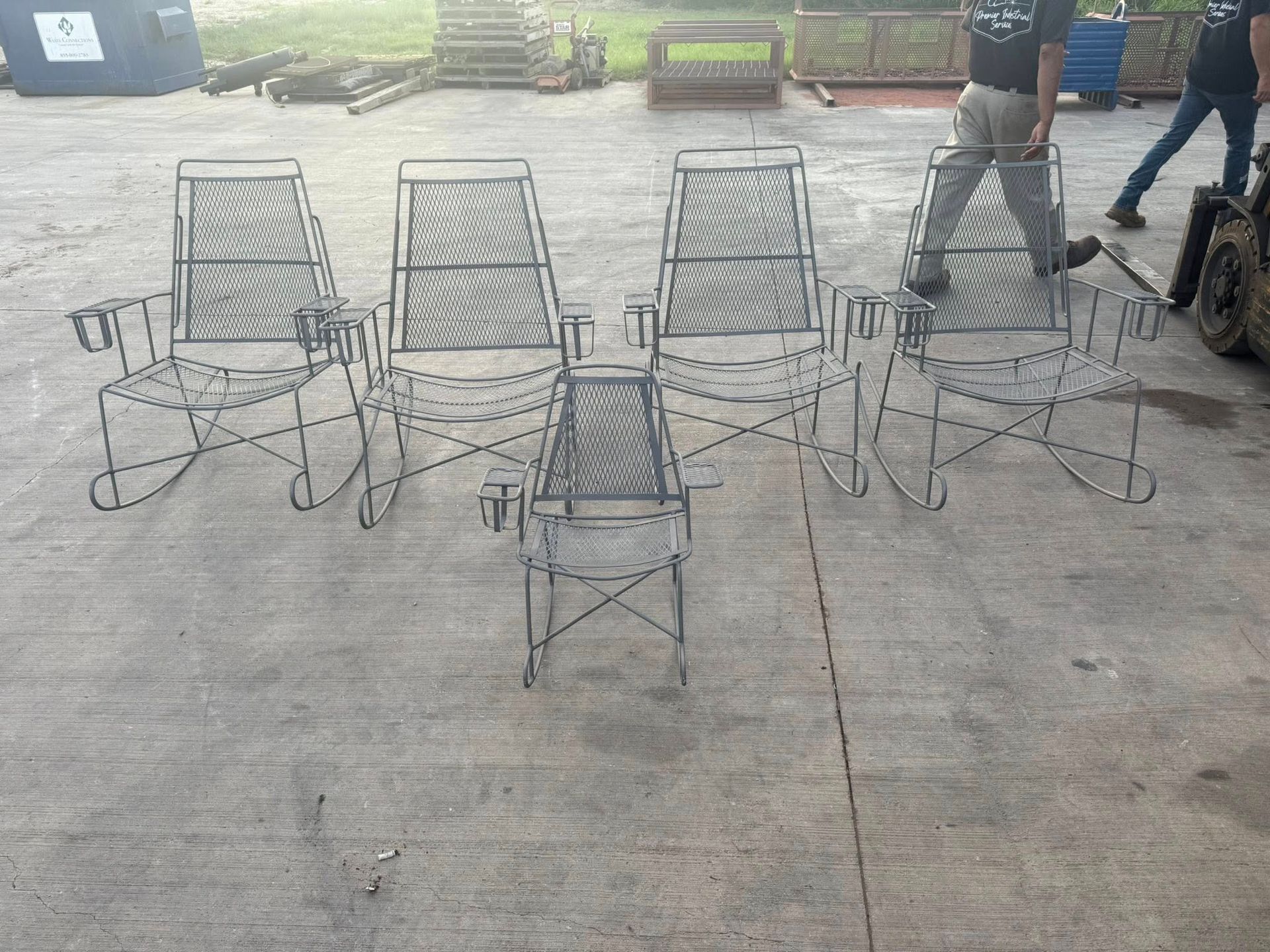 Four metal rocking chairs and a small grill sit on concrete. A person walks near the chairs.