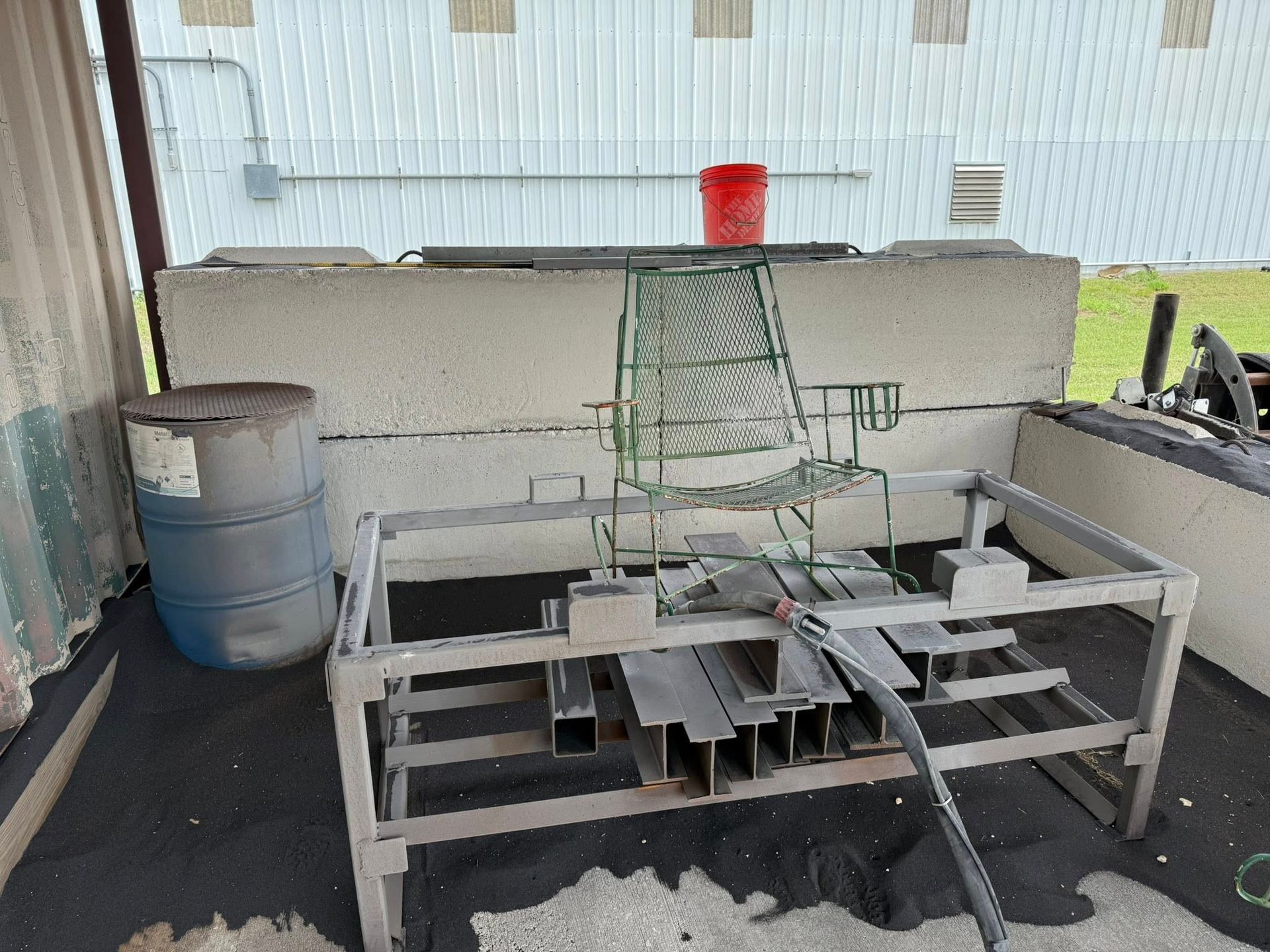 Outdoor industrial area with metal rack, barrel, and green metalwork against a building.