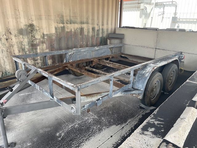 Gray, rusty utility trailer with two wheels, in front of a corrugated wall.