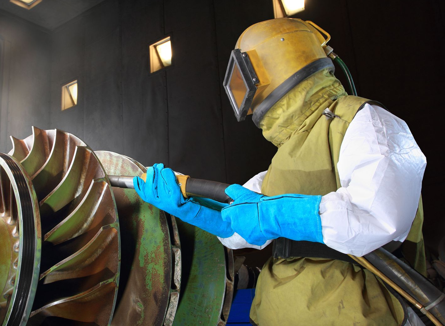 Person wearing protective gear sandblasting a large metal turbine.