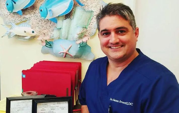 A man in a blue scrub is smiling in front of a painting of fish on a wall.