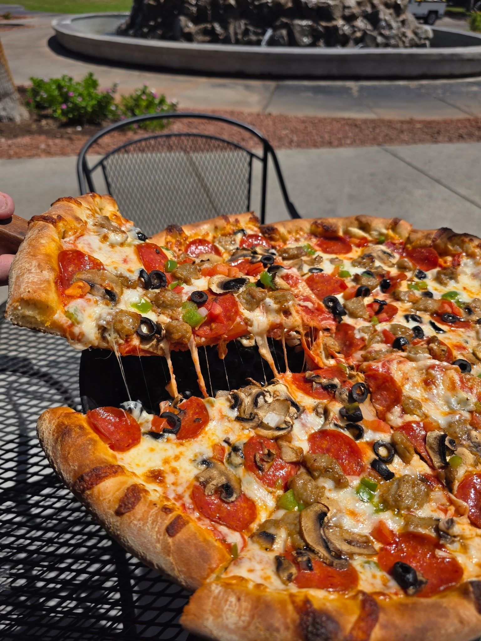 Pizza slice with melting cheese, pepperoni, mushrooms, olives and sausage, held over the table outside.
