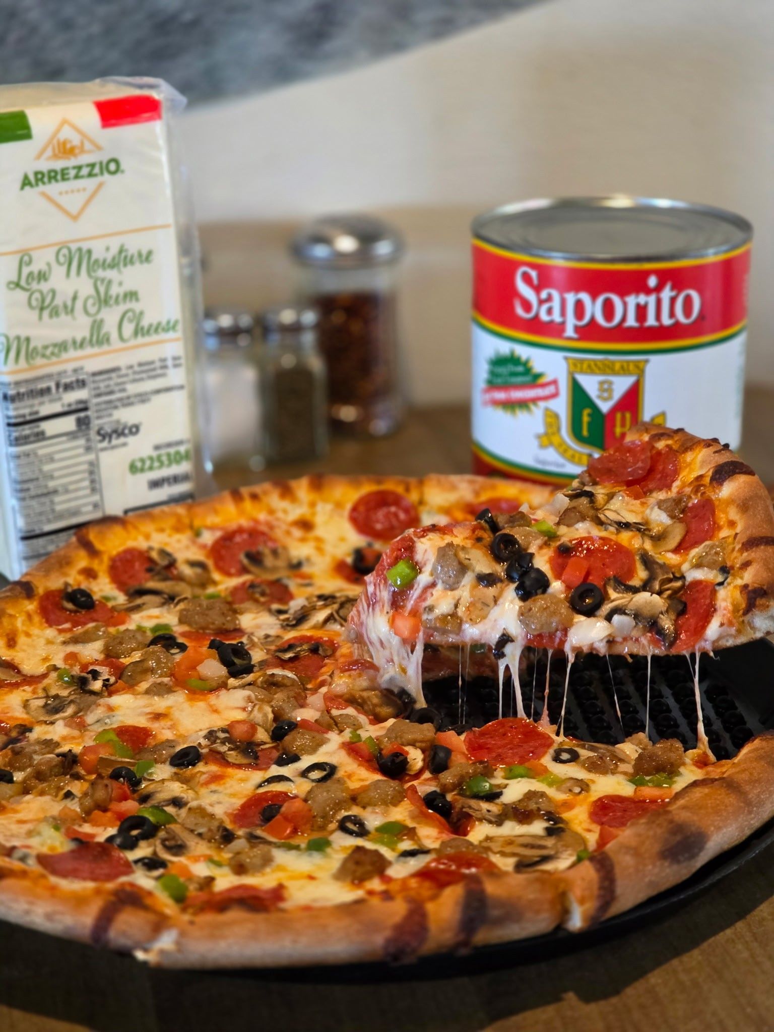Pizza with toppings, slice being lifted; cheese, sauce, cheese and seasoning ingredients nearby.