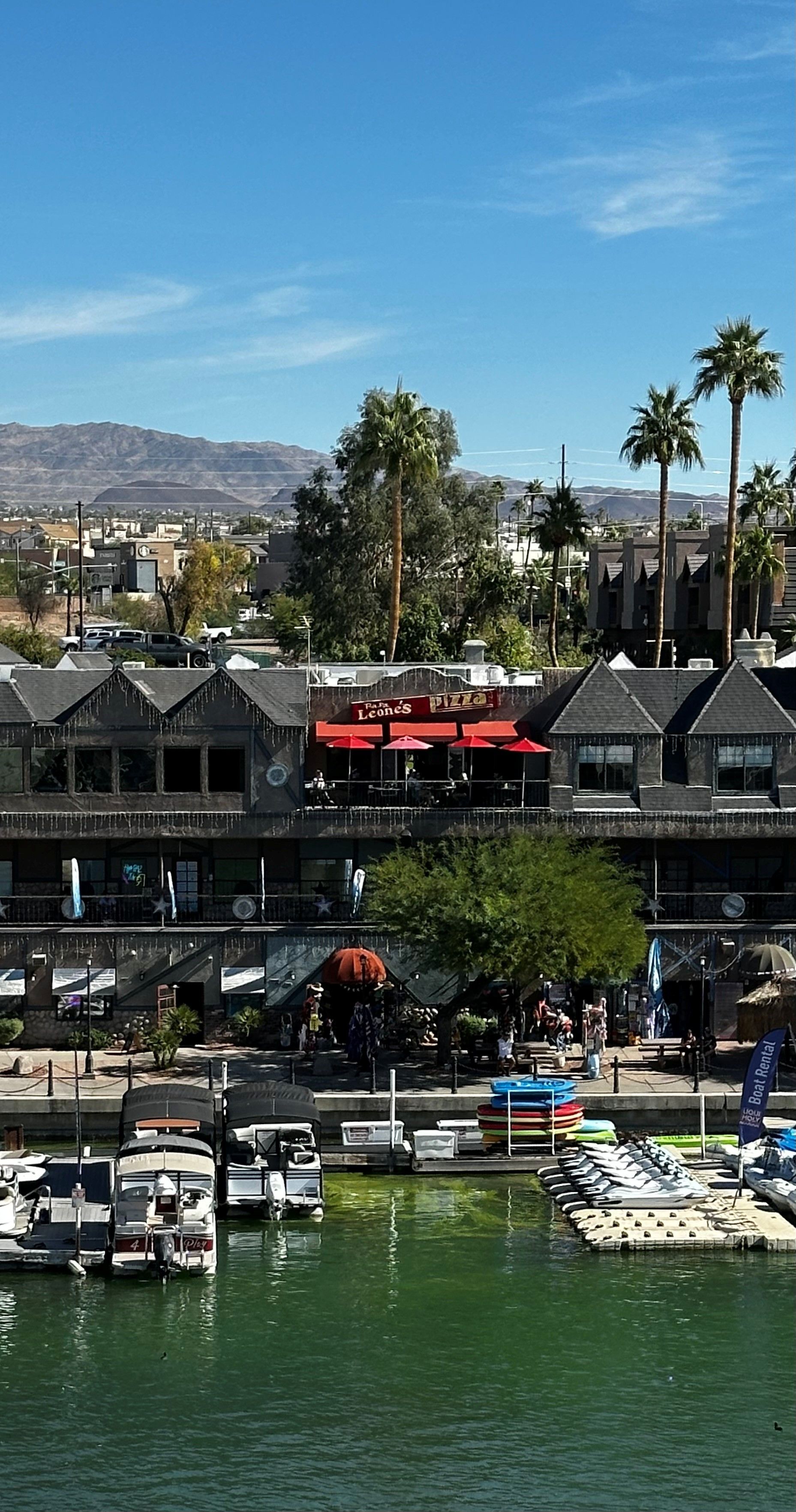 Boats docked on a lake, with buildings and a restaurant under a bright blue sky. Palm trees and mountains in background.