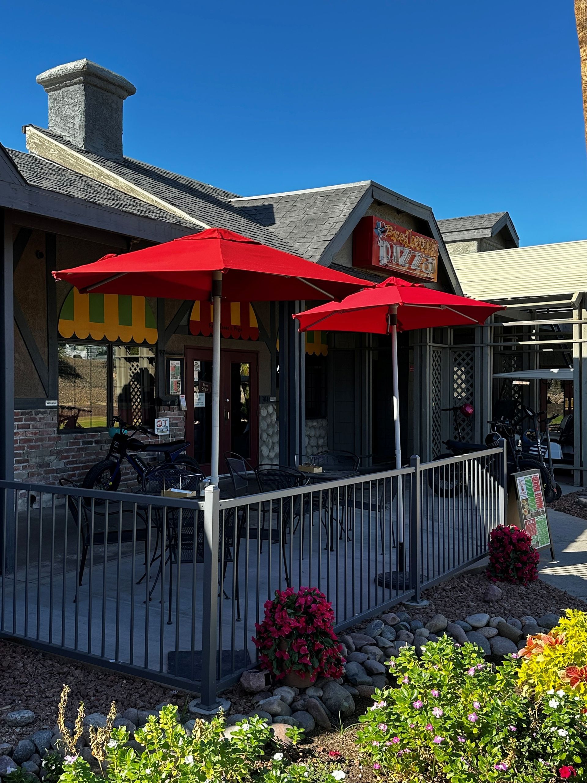 Restaurant patio with red umbrellas and small flowerbeds under a bright blue sky.