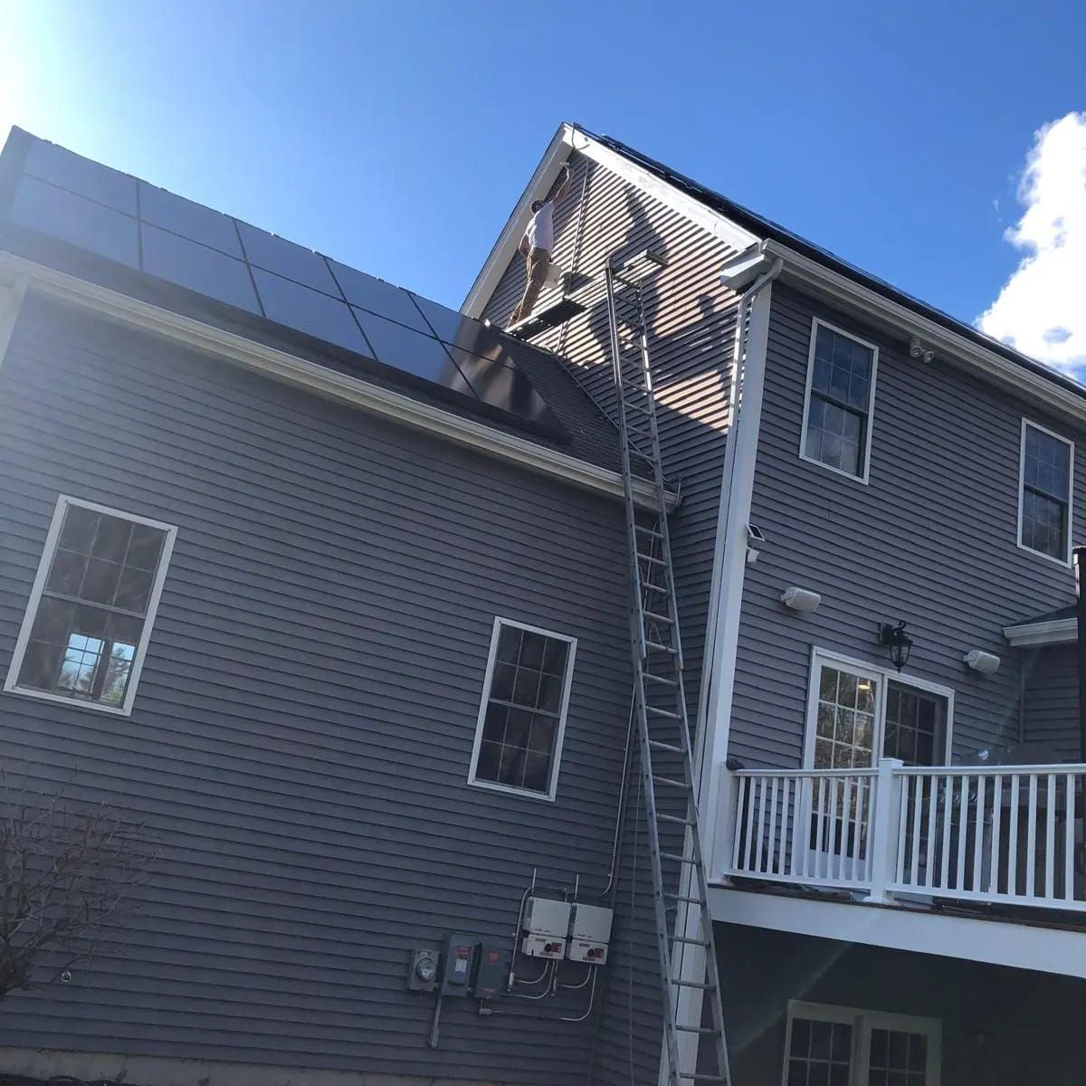 Two-story gray house with solar panels on roof; a tall ladder leans against the siding.