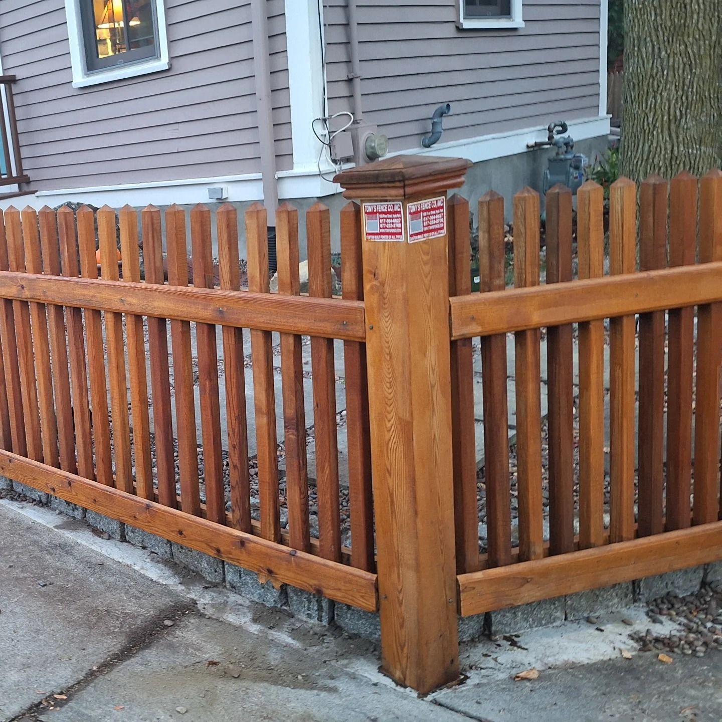 Wooden fence with a brown finish, cornering a sidewalk, next to a house with siding.