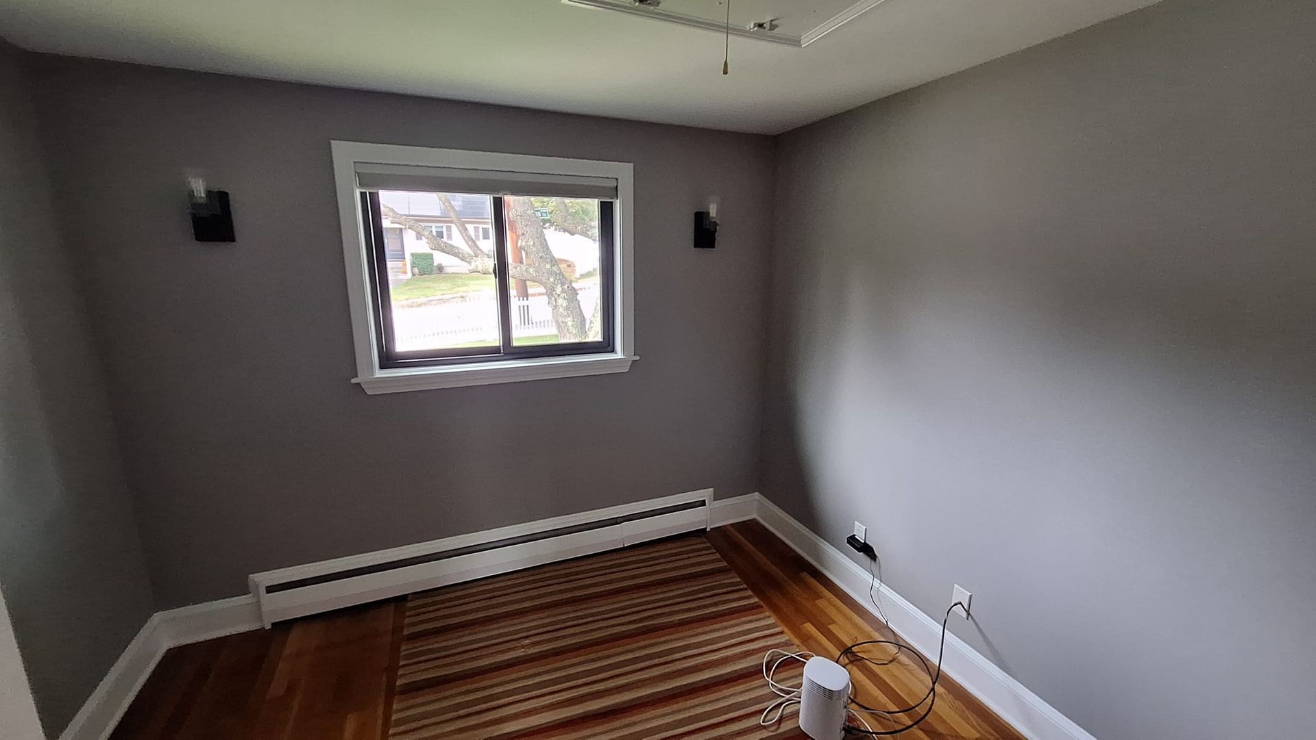 Empty room with grey walls, window, hardwood floor, baseboard heater, sconces, and overhead lighting.