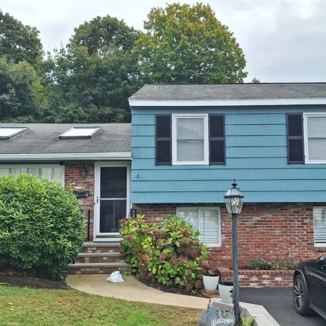 House with blue siding, brick, and black shutters, with bushes, trees, and a walkway.