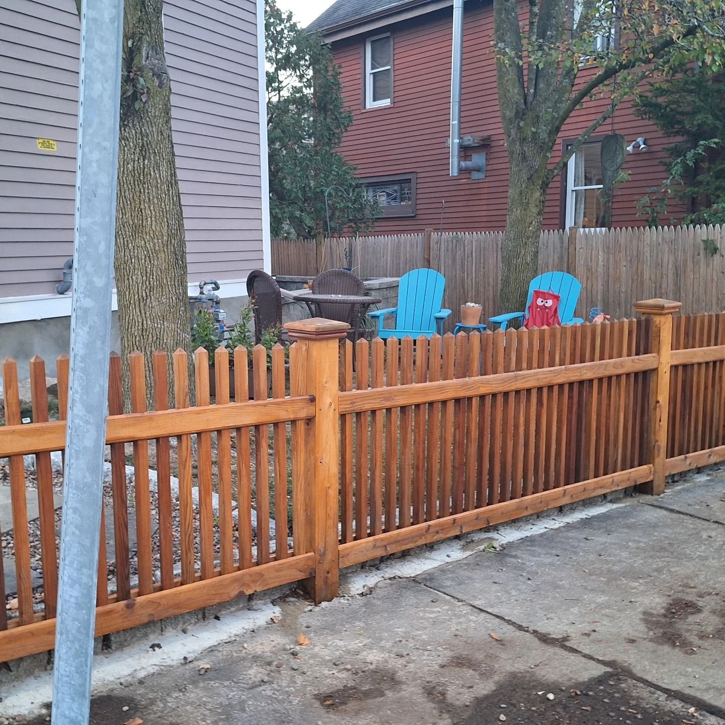 Wooden picket fence along a sidewalk, with colorful chairs visible in the backyard.
