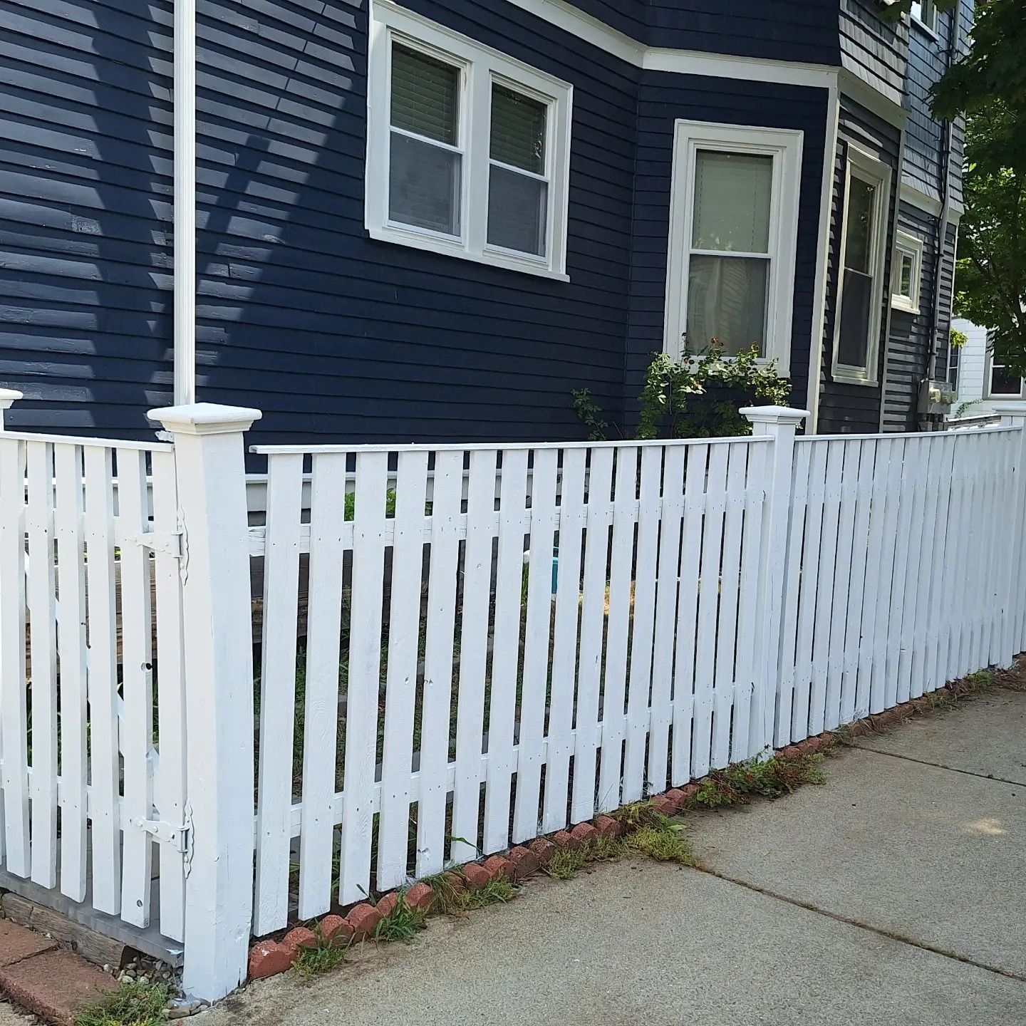 White picket fence in front of a blue house with windows, sidewalk on right.