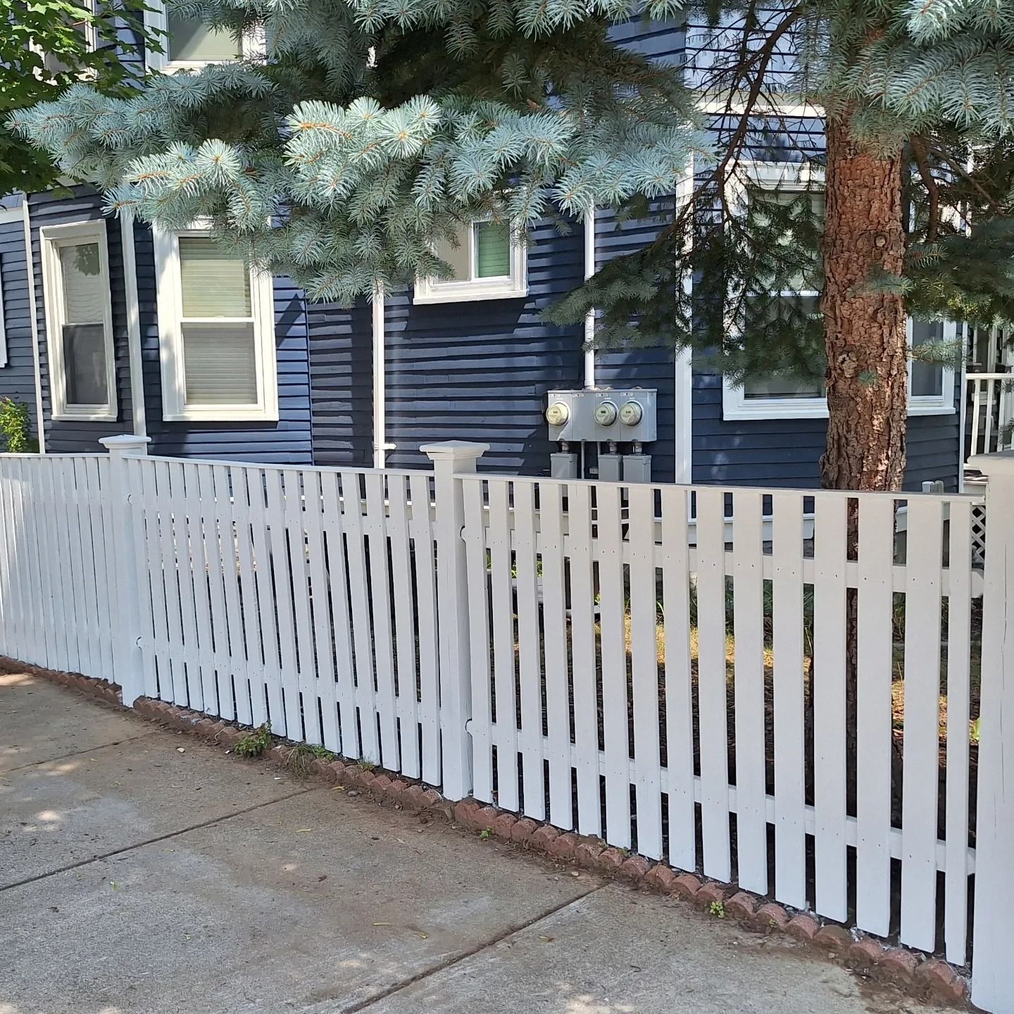 White picket fence in front of a blue house with trees overhead, near a sidewalk.