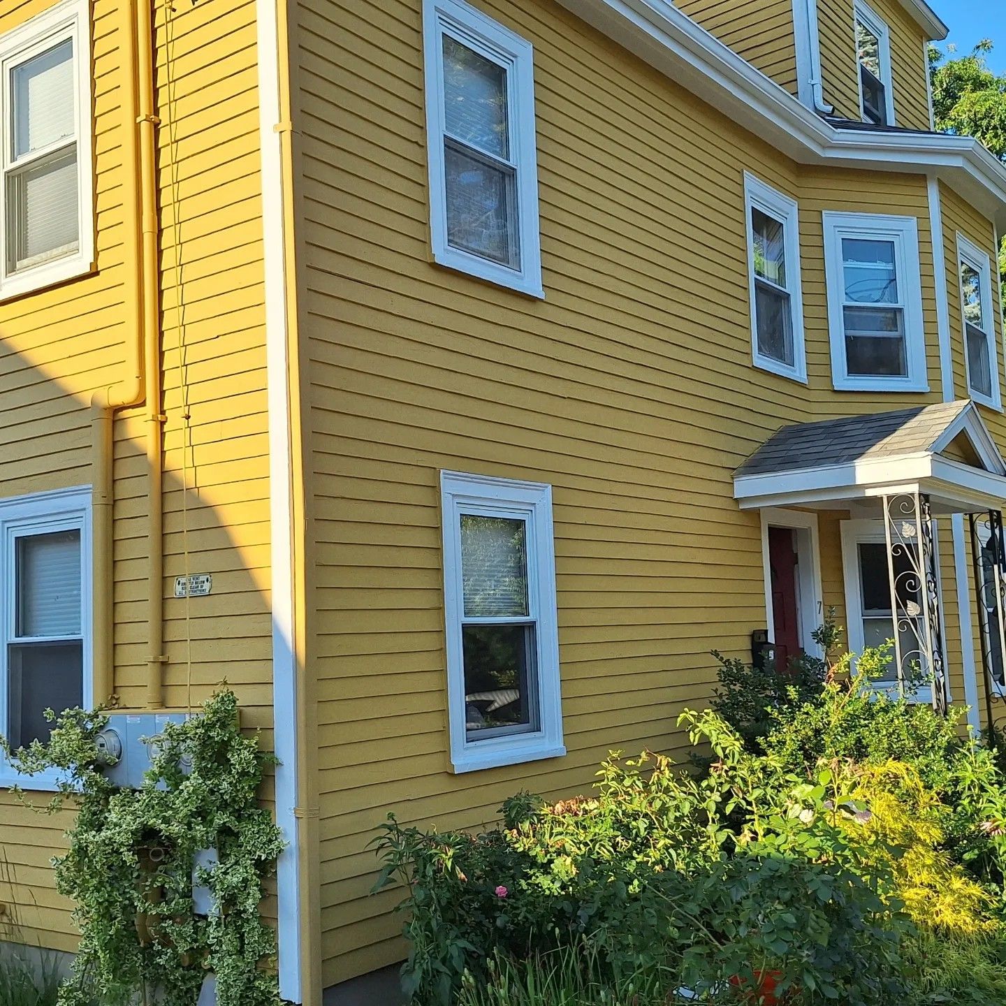 Yellow house with white trim, multiple windows, and a small front porch, surrounded by green bushes.