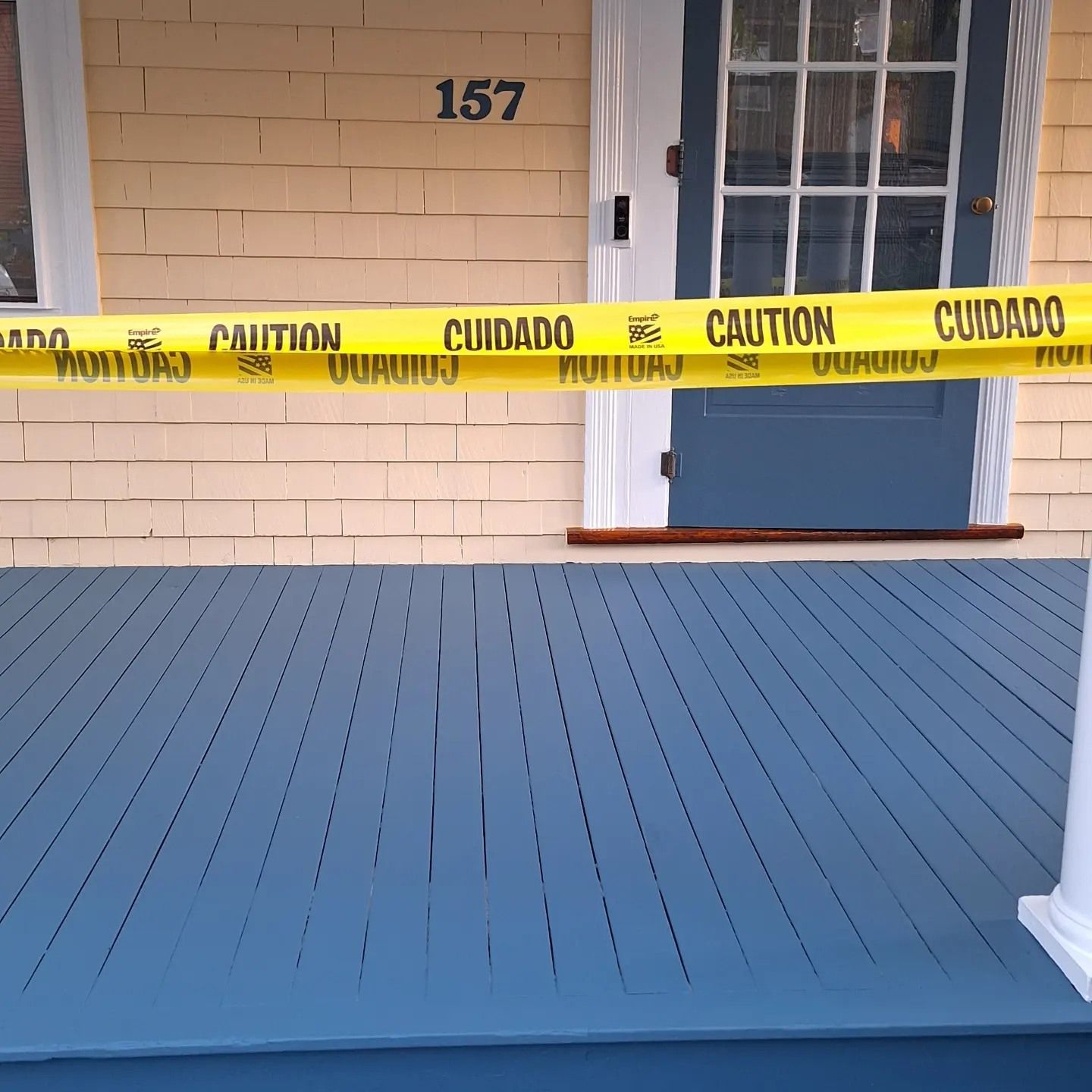 Yellow caution tape across a blue porch, in front of a blue door and a building with the number 157.