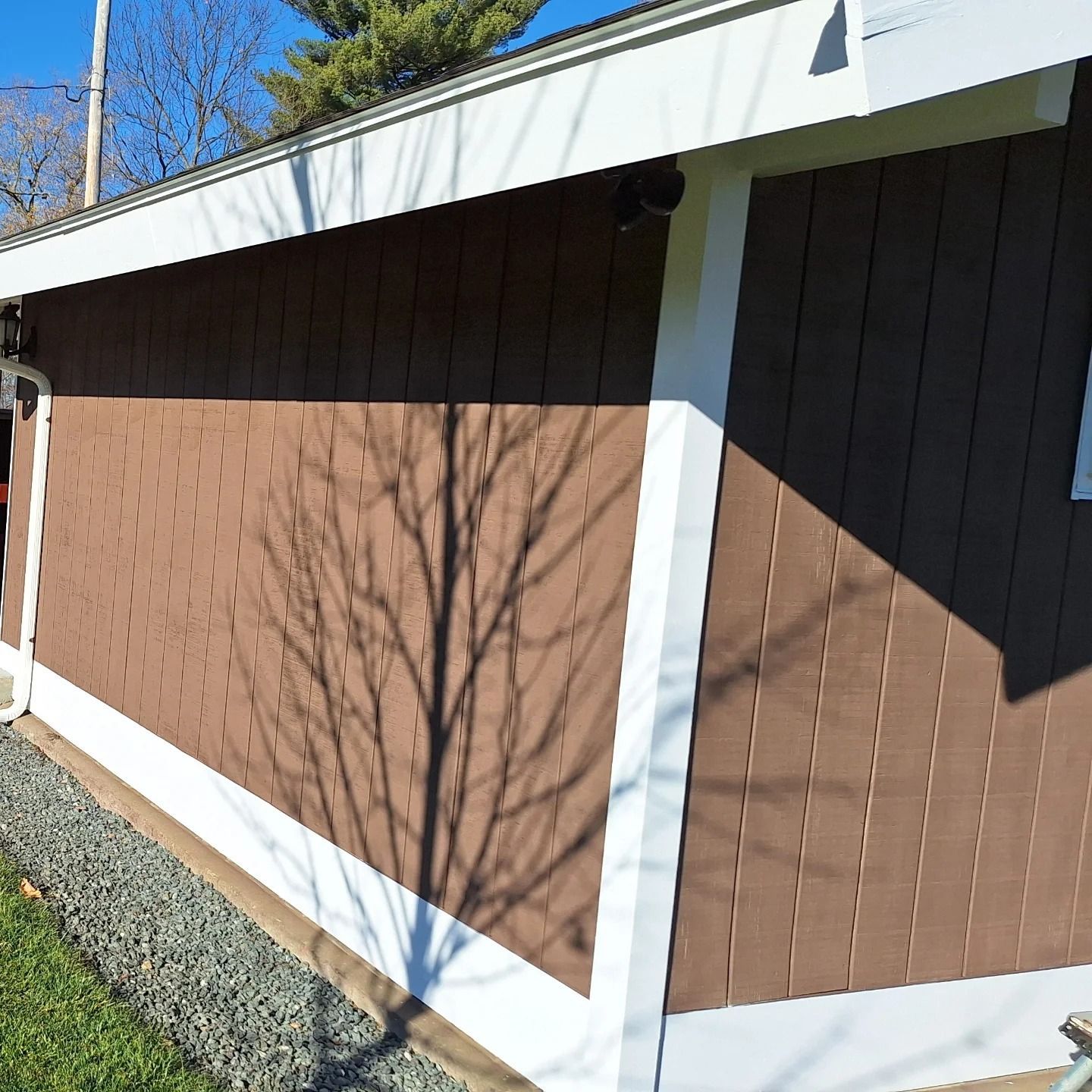 Brown building with white trim; tree shadow on the wall, grass and gravel below.