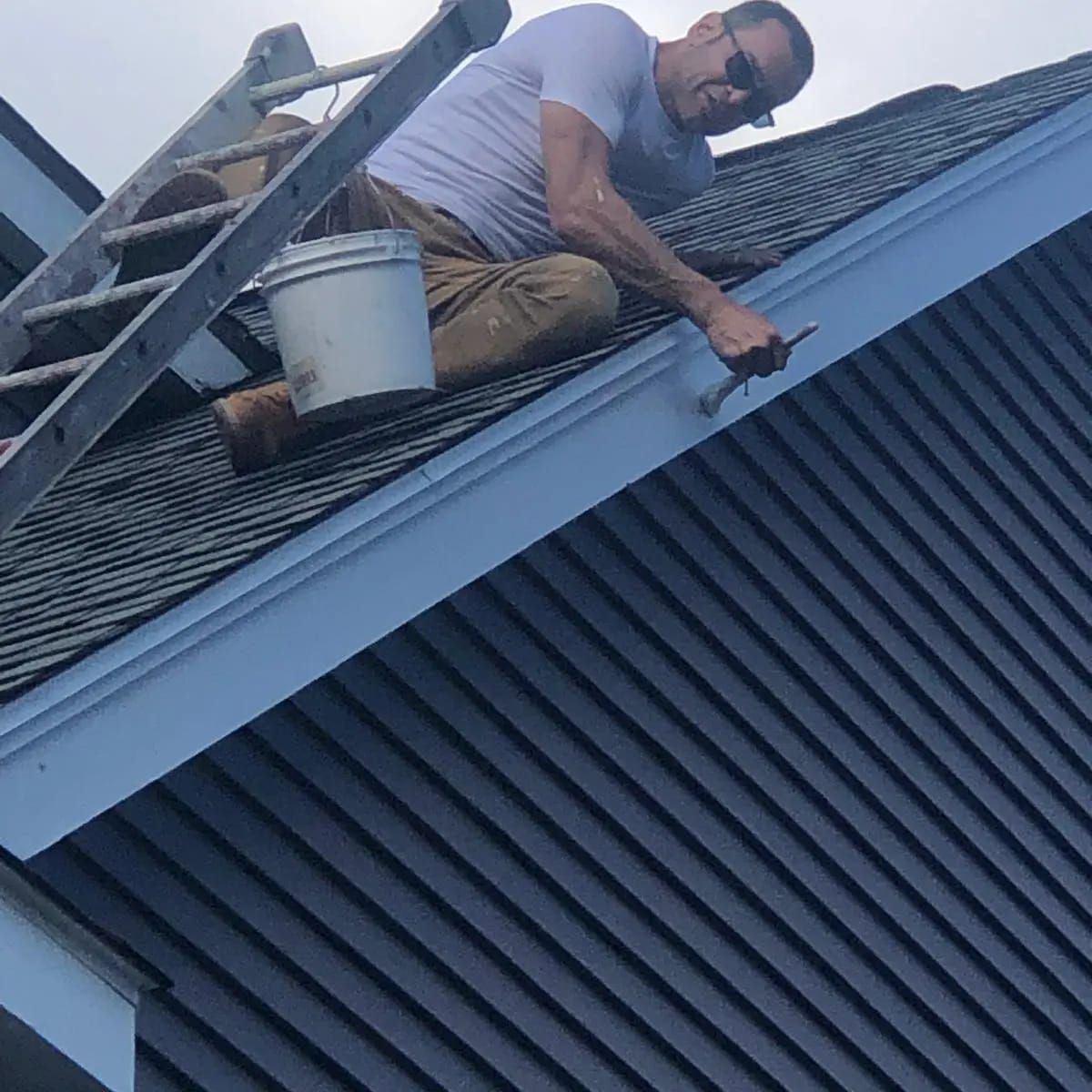 Man painting blue trim on a house roof, using a paintbrush, with a paint bucket nearby.