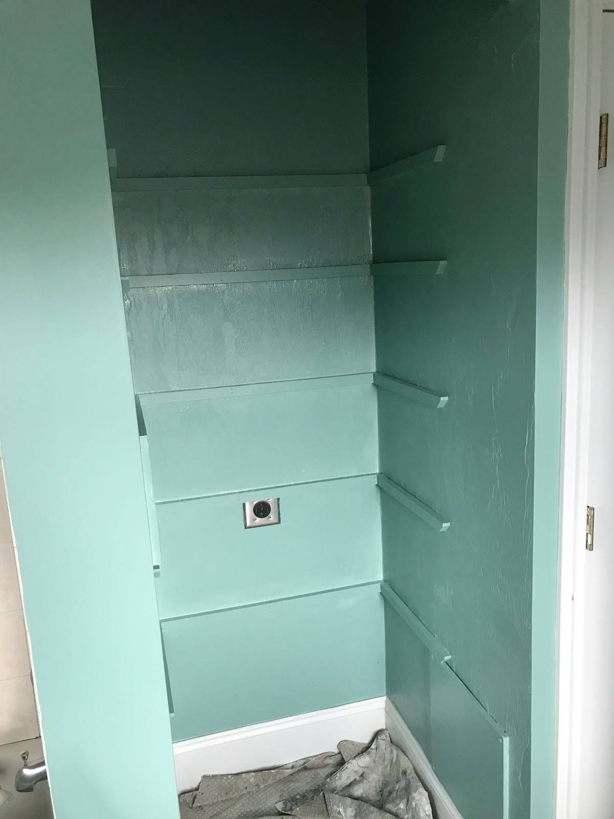 A built-in closet with shelves and brackets, painted aqua. A white baseboard and a light fixture are visible.