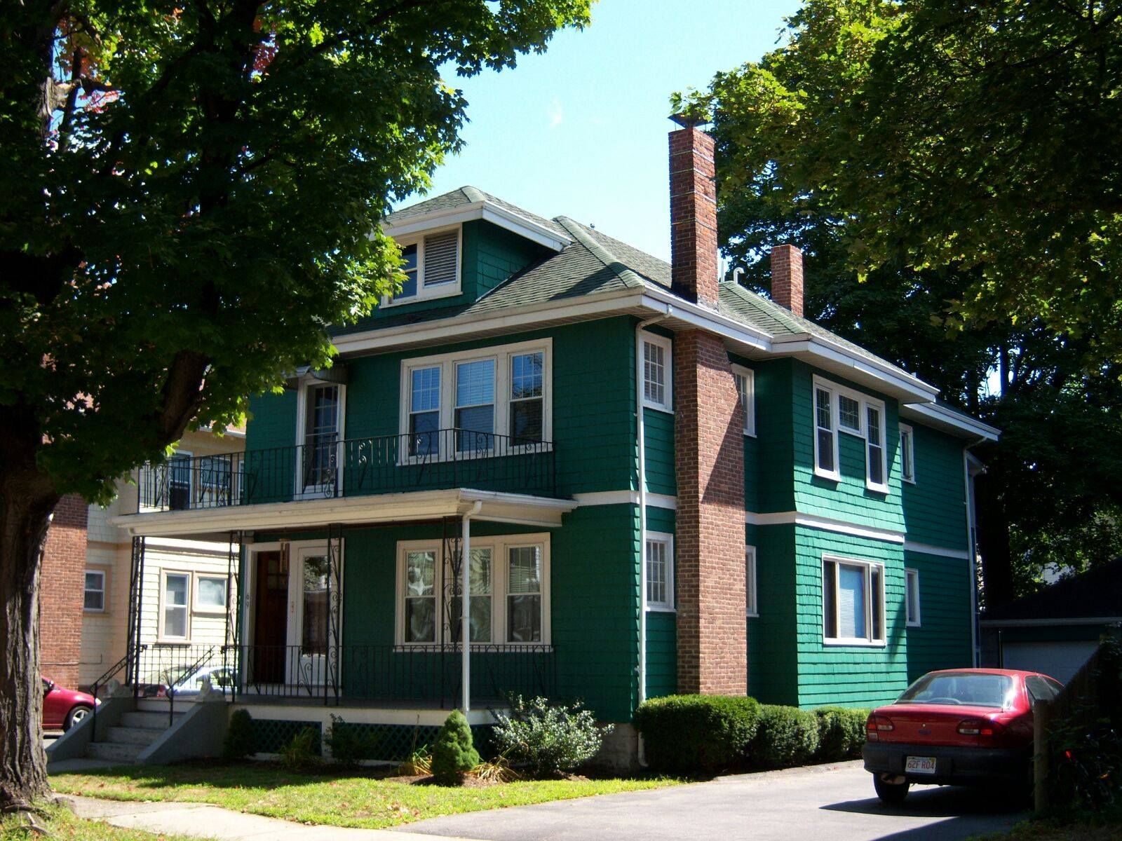 Two-story green house with a porch and brick chimney on a sunny day. A red car sits in the driveway.
