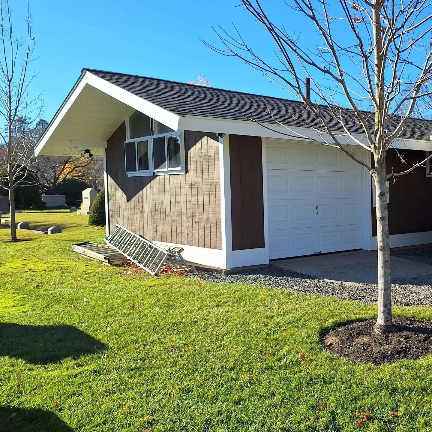 Garage with white door, brown siding, and dark roof on a sunny day. A ladder leans against the building.
