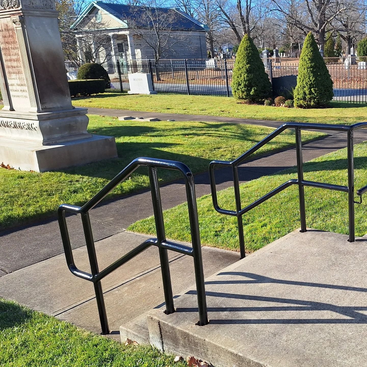 Black handrails on concrete steps in a grassy area with a cemetery and a house in the background.