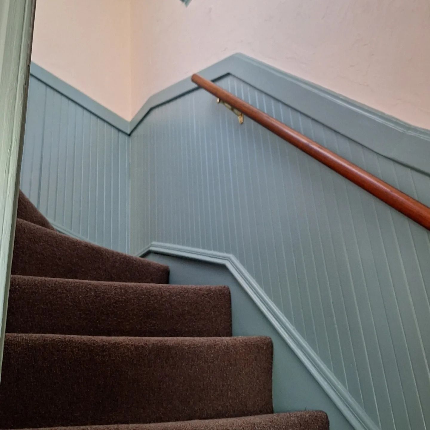 Staircase with brown carpet steps and a teal paneled wall with a wooden handrail.
