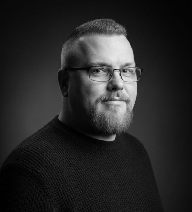 Black-and-white portrait of a bearded person wearing glasses and a dark sweater, looking at the camera