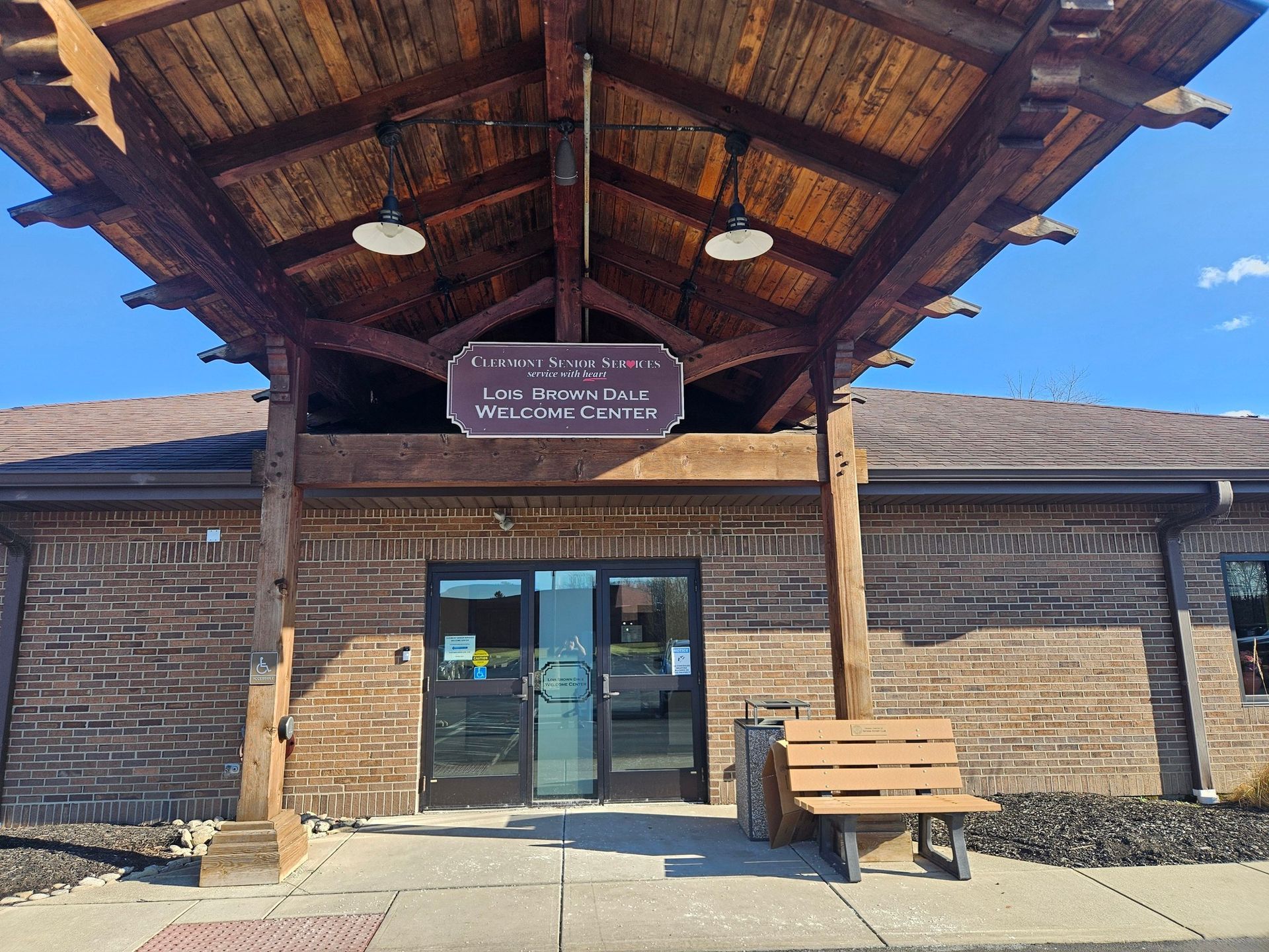 Rustic brick building entrance with wooden canopy, glass doors, sign, and bench in bright sunlight