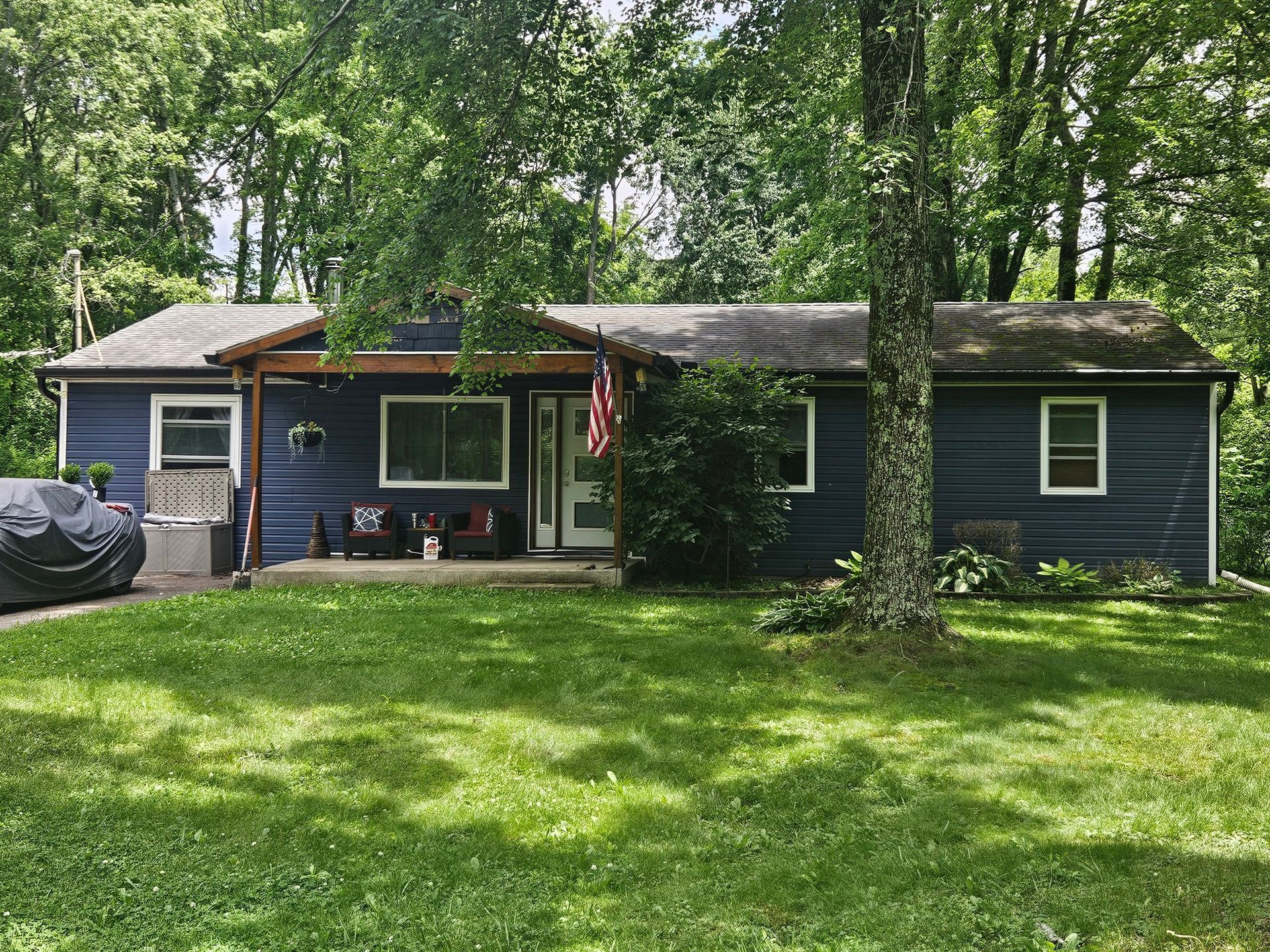 Small dark-green house with a porch and American flag, surrounded by trees and a grassy lawn.