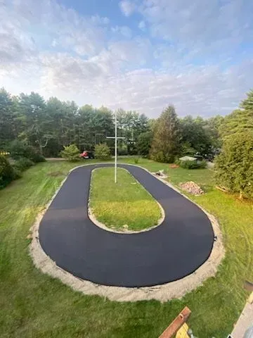 Oval asphalt driveway in grassy area, surrounded by trees and a cloudy sky.