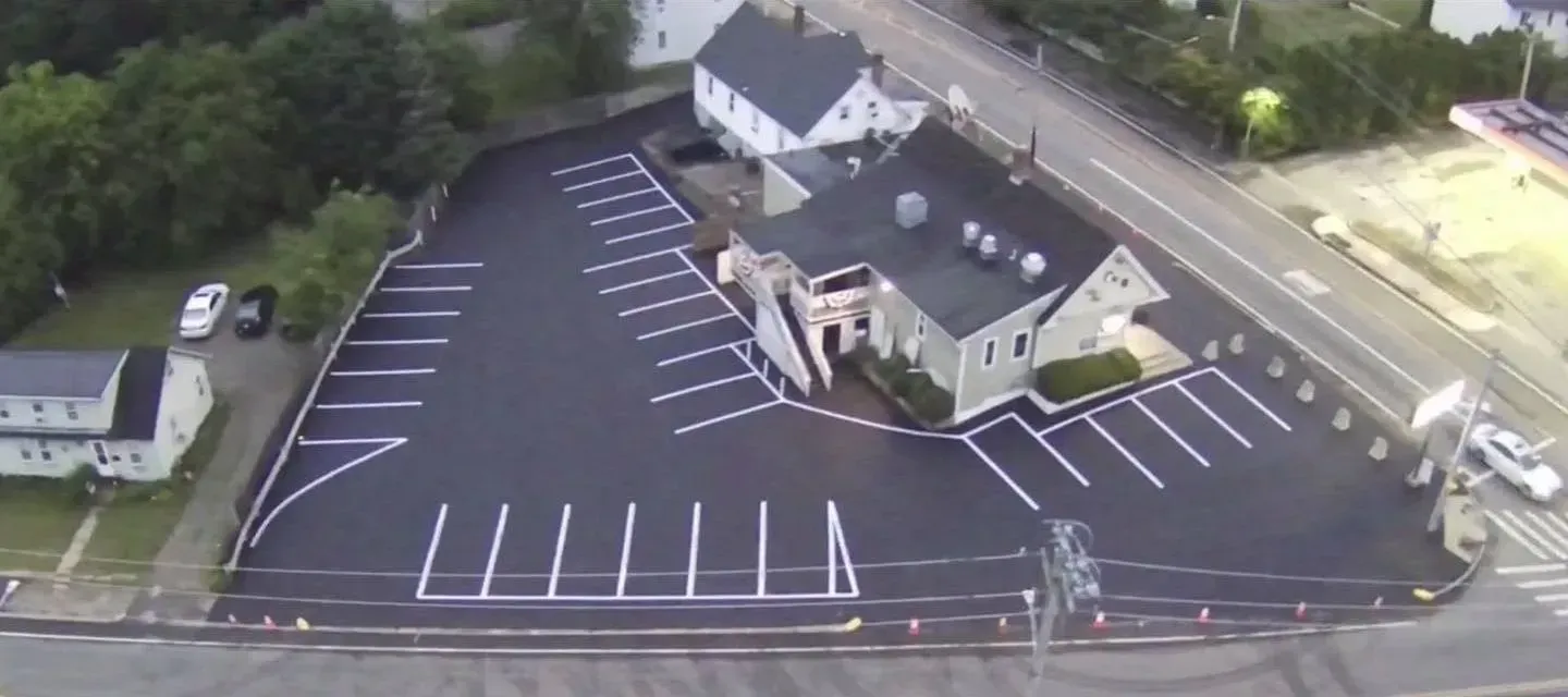 Aerial view of a parking lot next to a building and a road. The lot has marked parking spaces.
