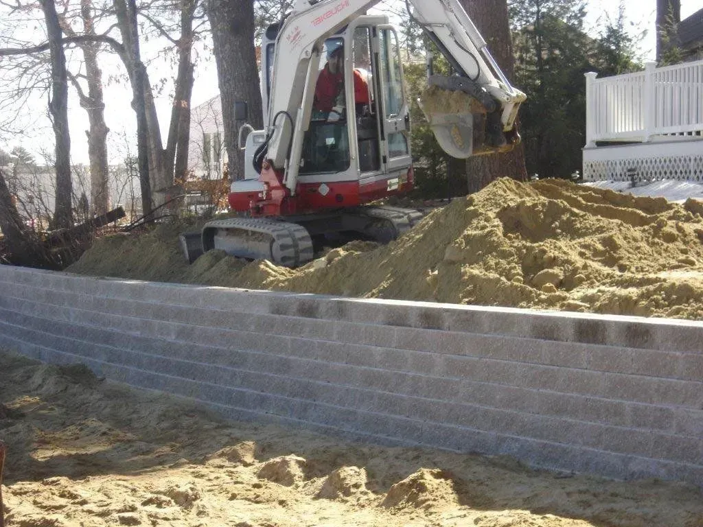 An excavator placing soil on a retaining wall made of gray blocks.