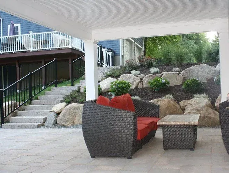 Patio with dark wicker furniture under a white covered area, next to stone steps and a rocky hillside.