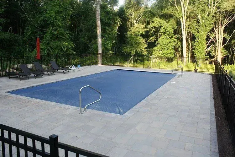 Swimming pool with a blue cover, surrounded by stone patio and black fence, wooded area in background.