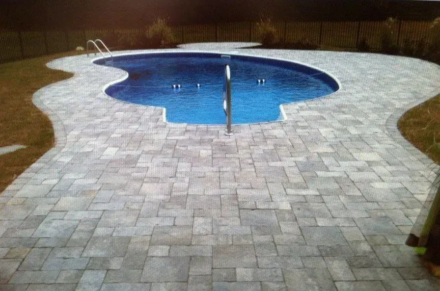 A blue kidney-shaped pool surrounded by grey pavers and grass, with a dark fence in the background.