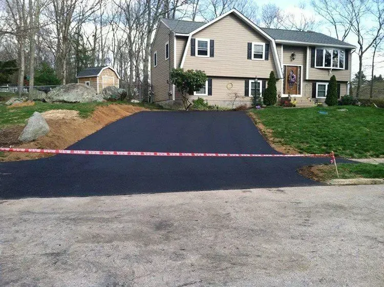 Newly paved black asphalt driveway in front of a tan house with a bay window and dark roof. Red caution tape across driveway.