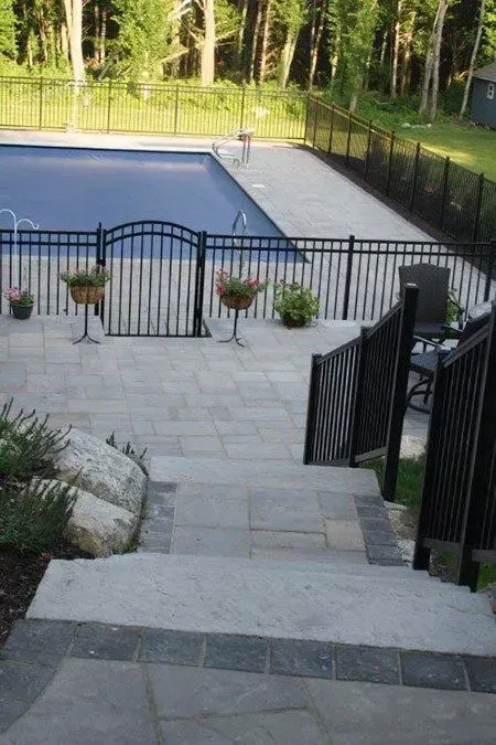 Stone steps lead down to a paved patio, pool, and black metal fencing in a yard with trees.