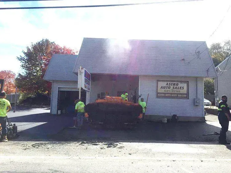 Paving crew paving asphalt in front of Astro Auto Sales, a small business building with open garage doors.