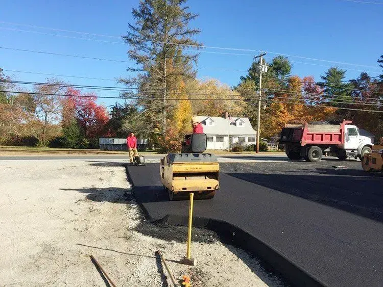 Asphalt paving in progress: Roller compacts fresh blacktop. Workers and dump truck present on a sunny day.