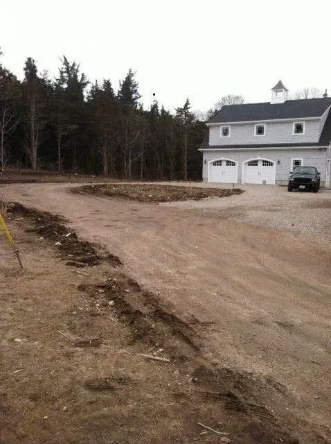 Dirt driveway leading to a two-story gray house with a two-car garage; trees in the background.