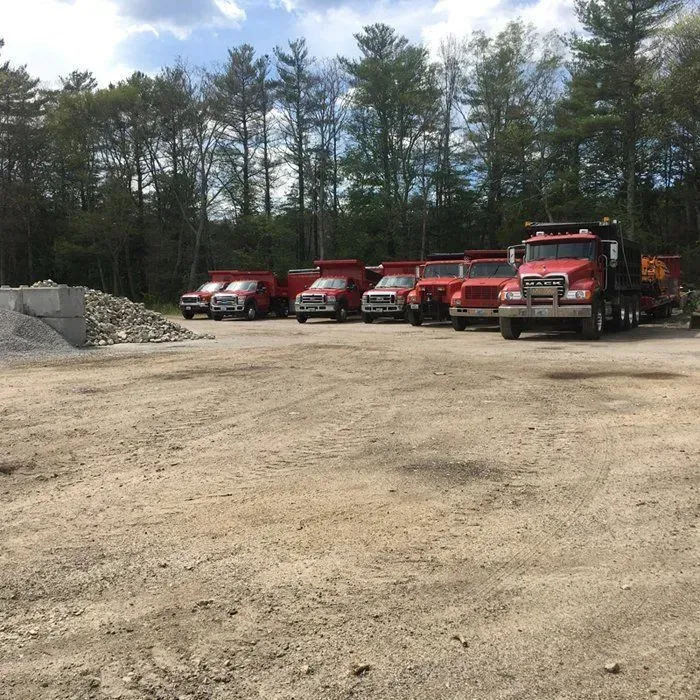Red dump trucks parked on a dirt lot with a pile of gravel and trees in the background.