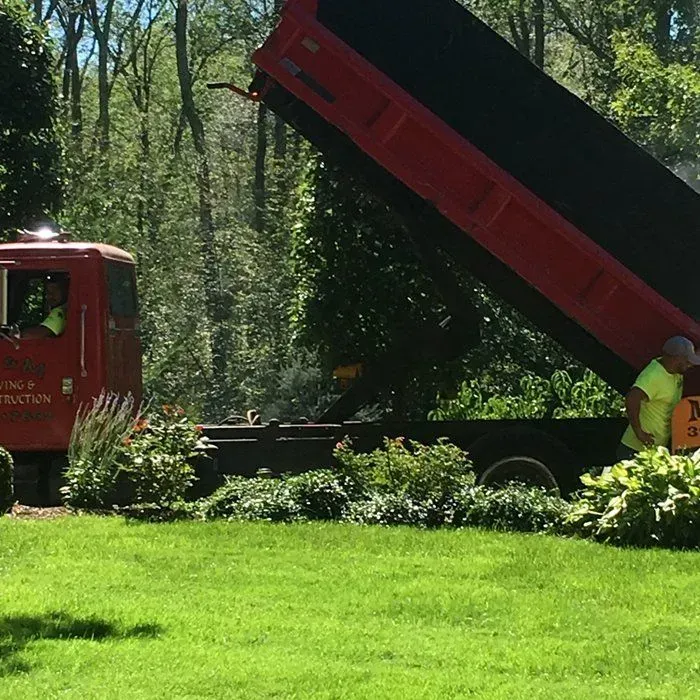 Red dump truck unloading in a yard with workers. Green grass, trees in background, sunny.