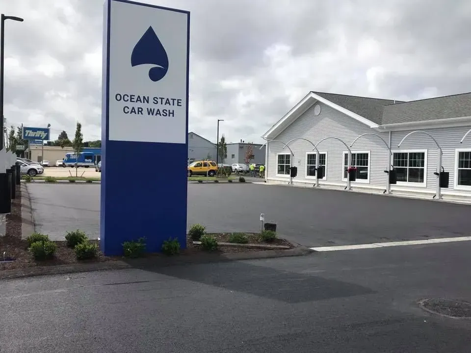 Ocean State Car Wash sign in front of a gray building on a cloudy day. Blue sign with logo.