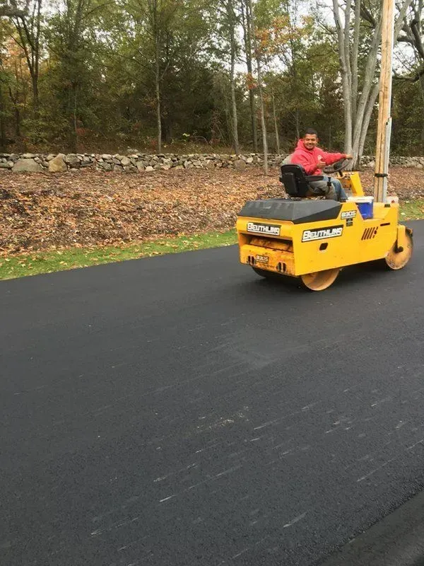Person operating a yellow asphalt roller on a freshly paved road. Trees in background.