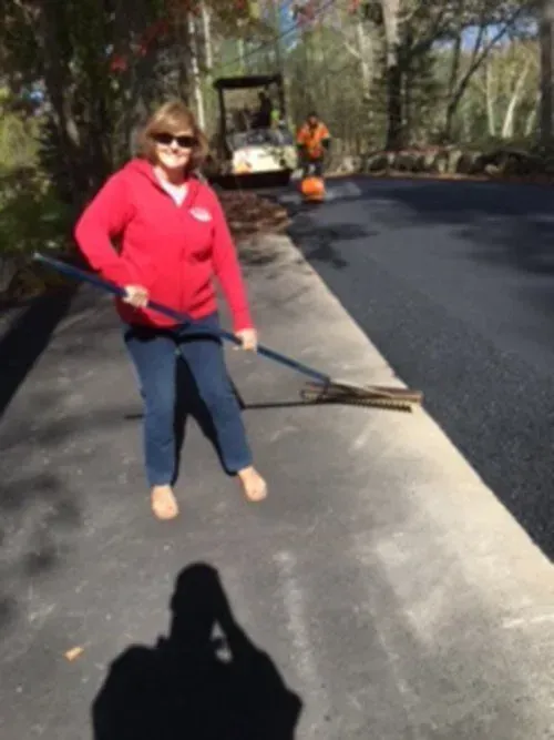 Woman in red sweatshirt uses a rake on fresh asphalt. Another worker and machinery are visible in the background.