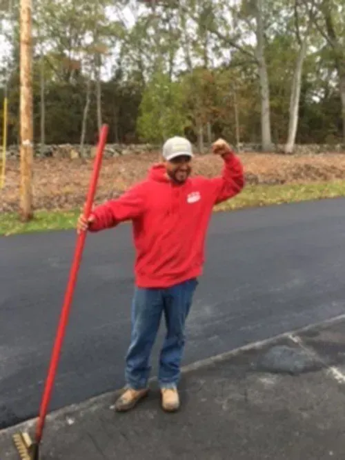 Man in red hoodie flexing, holding a rake on a paved road, smiling.
